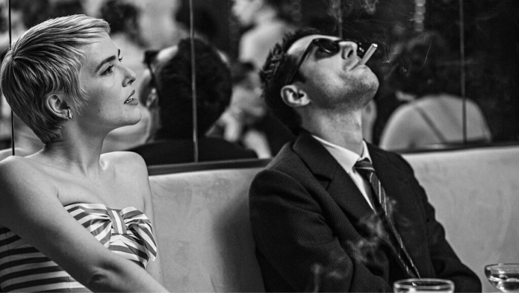 Guillaume Marbeck i rolla som regissør Jean-Luc Godard, iført dress, svarte solbriller og med ein sigarett i munnen i lag med Zoey Deutch, frå filmen Nouvelle Vague (2025).